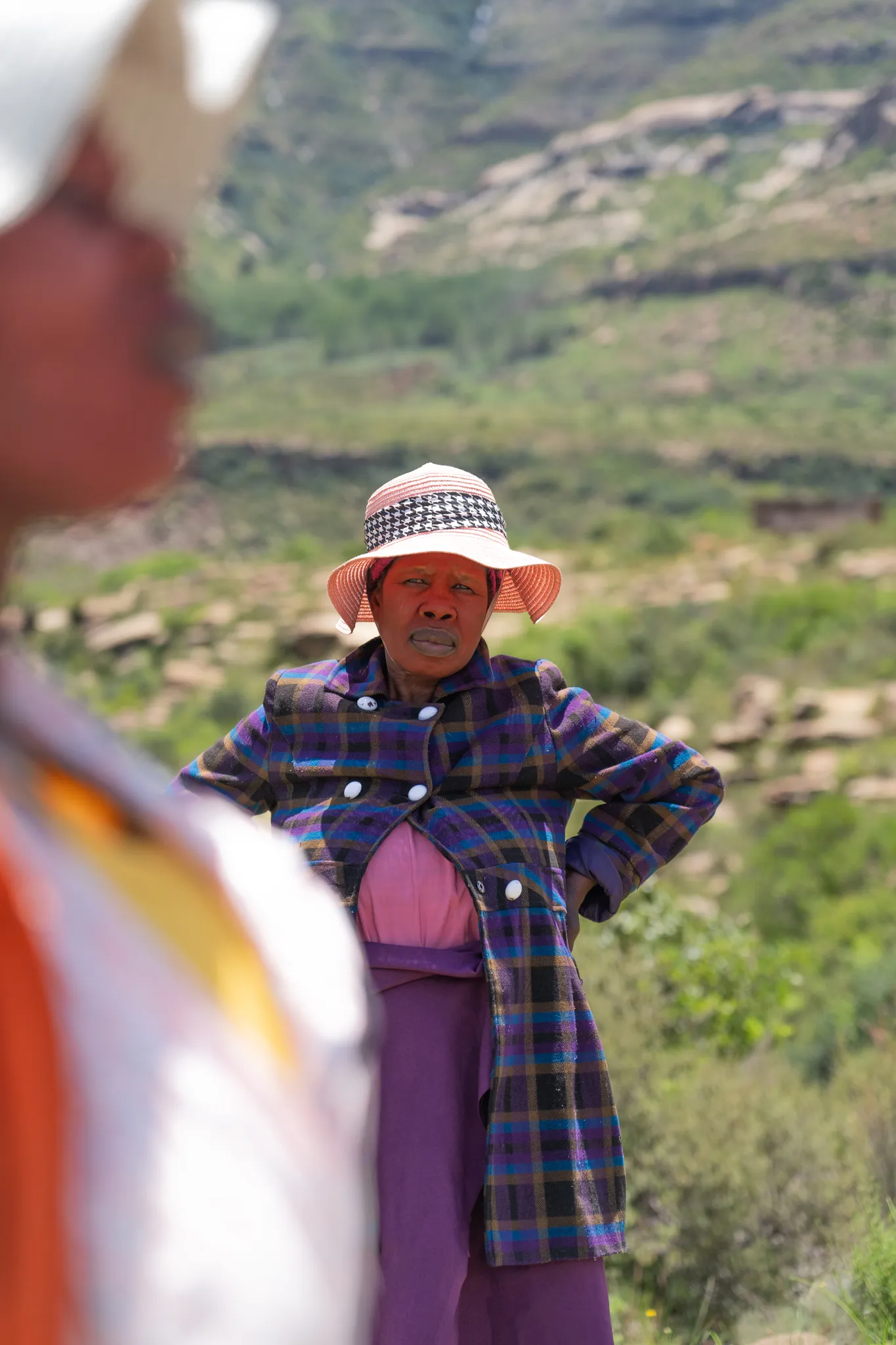 Woman in plaid coat and straw hat, hands on hips, Lesotho